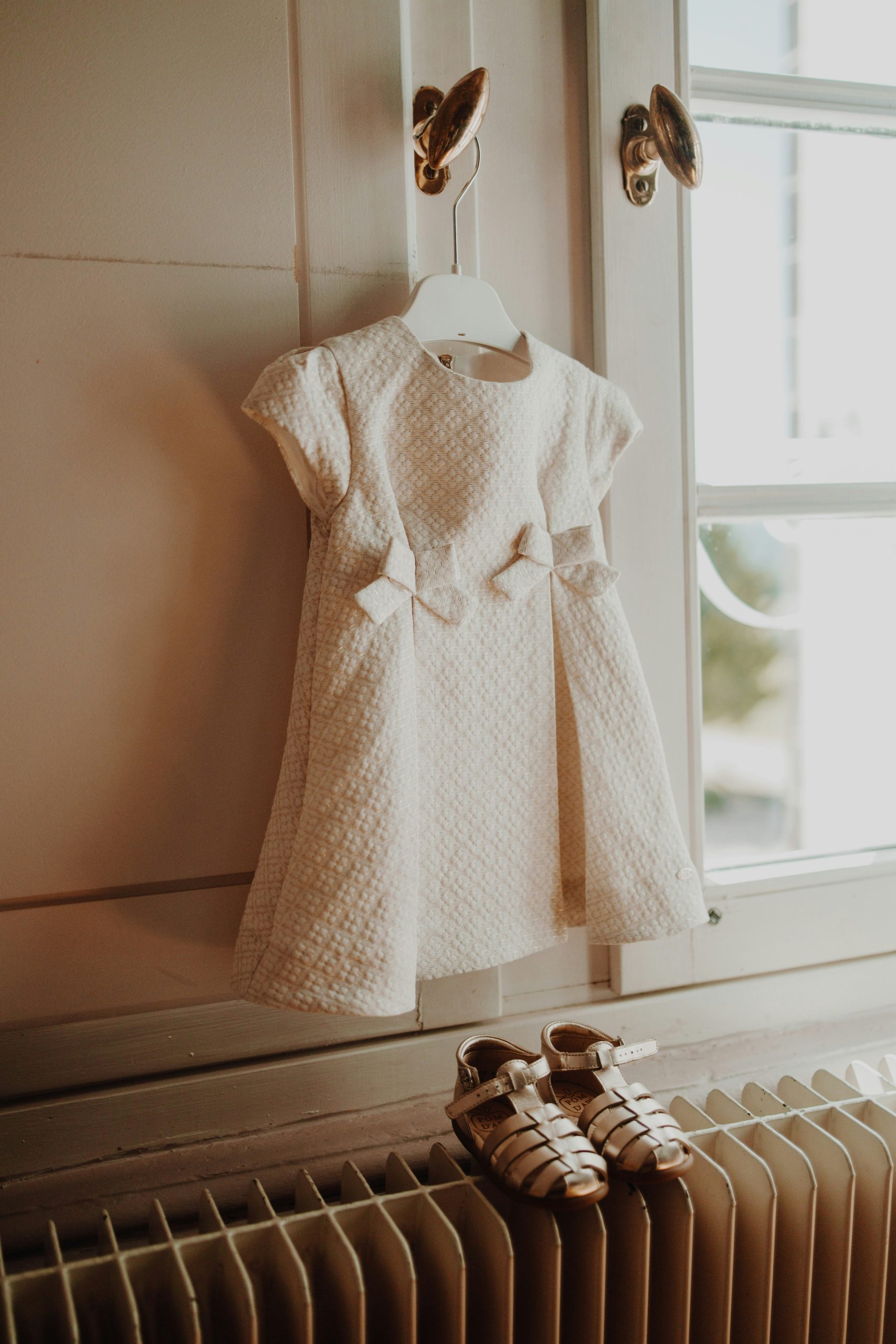 Cream coloured baby dress hanging on a door hook with small matching shoes placed below near a window.