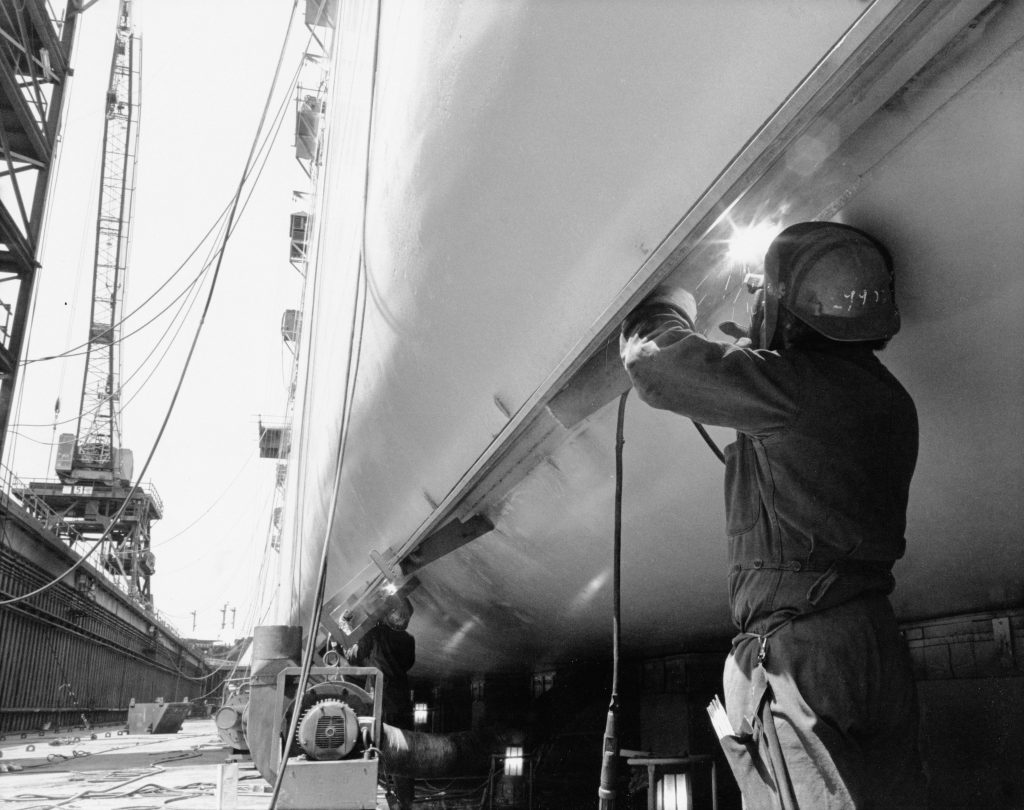 Welding operations on a large ship hull supporting shipyard supply chain management and sourcing operations. A skilled welder performs structural welding on a large steel ship hull, highlighting controlled fabrication processes that support shipyard supply chain management, procurement logistics, and reliable sourcing operations for marine vessels.